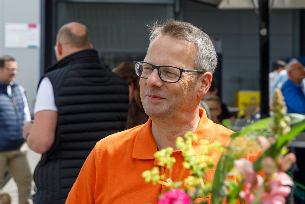 Mann (Mitarbeiter) in einem orangen T-Shirt beim Sommerfest vom MEDIAHAUS. Unscharf im Hintergrund weitere Mitarbeiter.