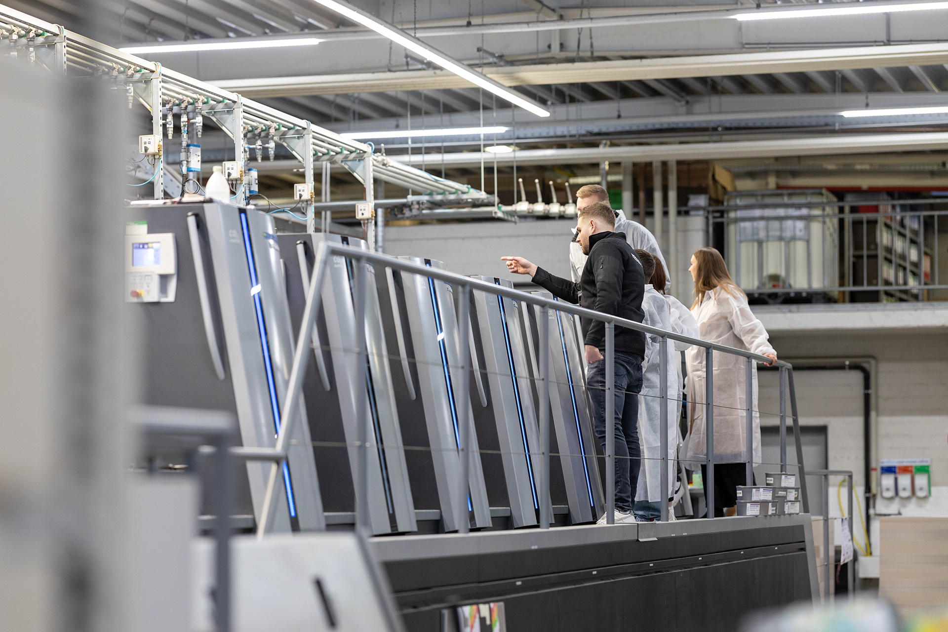 Druckereiführung - Mitarbeiter, der den Besuchern in Schutzkleidung die Druckmaschine zeigt.