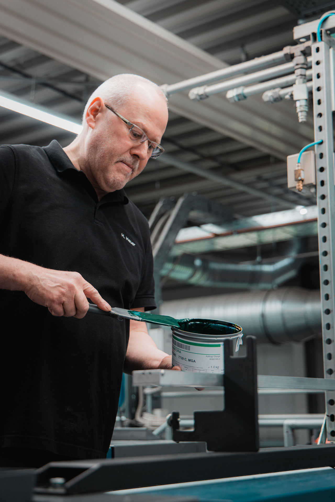 Druckerei-Mitarbeiter mit einem Farbeimer mit grüner Farbe in der Hand an einer Druckmaschine.